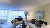 This single-floor space centers around a thoughtfully designed living room that serves as the focal point of the home. The room is characterized by large floor-to-ceiling windows dressed with vertical blinds, allowing abundant natural light to fill the space while offering the option for privacy. The pale, neutral-toned walls complement the light carpet flooring, contributing to an open and airy ambiance throughout.

The living room is furnished with two generously sized cream-colored sofas, each accented with decorative pillows in neutral and dark tones, creating a cohesive and stylish aesthetic. A round white coffee table anchors the seating arrangement, topped with a decorative tray and candle holders. A tall indoor plant adds a natural element to the corner near the windows, enhancing the room's freshness.

Wall décor includes a large framed artwork depicting a bridge scene, adding visual interest to the space. Warm ambient lighting from a classic table lamp provides a welcoming glow during evening hours. Overall, the living area exudes a clean, contemporary style with a comfortable and well-organized layout.