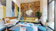 Located in the South End neighborhood of Charlotte, NC, this single-floor lobby space makes a striking first impression with its thoughtfully designed interiors and energetic aesthetic. The main lounge area features soaring ceilings and polished concrete floors that amplify the sense of openness. A dramatic floor-to-ceiling reclaimed wood accent wall, arranged in a geometric chevron pattern, anchors the seating area and provides a warm, textural contrast to the crisp white walls. A large circular mirror is mounted at its center, reflecting the stylish pendant lighting above. The lounge is furnished with a mustard yellow velvet sofa, contemporary lounge chairs, a marble-topped coffee table, and teal upholstered ottomans, creating a welcoming and colorful gathering space. Opposite the lounge, a bold, large-scale city mural depicting the Charlotte skyline commands an entire wall, infusing the space with local character and artistic flair. Floor-to-ceiling glass entry doors flood the lobby with natural light and offer clear views of the surrounding streetscape. A dedicated mail room features illuminated black parcel lockers lining both walls, accented by ambient LED underlighting, a patterned rust-toned area rug, and an eclectic gallery wall of framed artwork, completing this well-appointed and visually dynamic lobby experience.