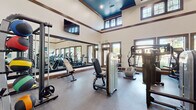 This fitness center is designed to support a full range of workout routines in a bright, open environment. The space features soaring ceilings with a striking teal-painted tray ceiling detail accented by recessed lighting and a ceiling fan, creating an airy and energizing atmosphere. Rich wood-trimmed moldings and framing throughout lend a refined, polished aesthetic to the space.

Large windows line the walls, flooding the interior with natural light and offering views of the surrounding landscaped grounds. Full-length mirrors along one wall visually expand the space and assist with form during exercise.

The fitness center is comprehensively outfitted with a wide variety of equipment. The cardio section includes multiple treadmills, elliptical trainers, stair climbers, and stationary cycling bikes, all positioned to take advantage of the outdoor views. The strength-training area features a selection of cable and resistance machines, a weight bench with free weights, and a medicine ball rack. A wall-mounted flat-screen television adds to the amenity. Durable rubber flooring throughout the main workout area provides safety and comfort underfoot. The space also includes a small storage and accessory station, ensuring everything needed for an effective workout is readily accessible.