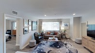 This single-floor space showcases a well-appointed living room designed with both comfort and style in mind. The room is anchored by a large picture window dressed with sheer curtains and vertical blinds, allowing an abundance of natural light to fill the space and offering views of the greenery outside. The light-toned walls and smooth white ceiling with recessed lighting contribute to a clean, airy atmosphere throughout.

The living area features rich hardwood-style flooring partially covered by a bold, large-scale floral area rug in navy and grey tones, adding a striking visual centerpiece to the room. Seating includes a neutral-toned sofa accented with colorful throw pillows in terracotta and teal, complemented by a sophisticated dark accent chair with a coordinating ottoman. A sleek glass-and-metal coffee table grounds the arrangement.

The walls are thoughtfully curated with an eclectic gallery of framed artwork in varied sizes, adding personality and depth to the space. Warm floor lamps and side tables with decorative accessories complete the polished, contemporary aesthetic, making this living space both functional and visually engaging.