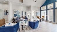 Located at 3415 SW 39th Blvd in Gainesville, Florida, this single-floor clubhouse is a thoughtfully designed communal space that blends modern aesthetics with welcoming functionality. The defining architectural feature is the soaring vaulted ceiling, which peaks at a dramatic height and is anchored by a bold cobalt blue accent wall. A large arched transom window and flanking French doors flood the space with abundant natural light, offering views of the outdoor pool area beyond.

The open floor plan flows seamlessly between distinct zones. The lounge area features plush navy velvet armchairs and a tufted gray sofa arranged around glass-topped coffee tables, all anchored by a large area rug. Large-scale abstract artwork in blue tones adds a cohesive, curated feel. Adjacent to the lounge, a spacious dining area centers on a solid wood farmhouse-style table with upholstered chairs, positioned beneath geometric pendant light fixtures and a wall-mounted television.

A dedicated café nook showcases a striking geometric patterned wallpaper accent wall, complemented by sleek white cabinetry and a coffee station. High-top bistro tables with cognac leather stools create a casual gathering spot. Light wood-tone flooring runs throughout, tying the entire space together with a clean, contemporary finish. A reception desk near the entrance completes the welcoming, well-appointed environment.