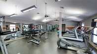 This fitness center is a comprehensive, single-floor facility designed to support a wide range of workout routines. The space is laid out efficiently, with distinct zones dedicated to cardiovascular training and strength work. Along one side, a row of treadmills and elliptical machines provides ample options for cardio exercise, while the opposite side houses a full free weights area complete with a tiered dumbbell rack, adjustable benches, and cable machines for targeted strength training. Large mirrors line the walls, visually expanding the space and allowing users to monitor their form throughout their workouts. The flooring features durable, rubberized material suited for heavy equipment and high-traffic use. Overhead, a combination of suspended fluorescent lighting and a ceiling fan ensures the space remains well-lit and ventilated. Vaulted ceiling sections add an open, airy quality to the environment. The neutral gray and warm orange accent color scheme gives the facility a clean, modern aesthetic. Overall, the fitness center is thoughtfully arranged to maximize usable floor space while offering a broad selection of equipment to accommodate users at varying fitness levels.