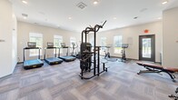 This well-equipped fitness center at Lodge View Drive in Rochester, Minnesota, offers a thoughtfully organized single-floor layout designed to support a full range of workout routines. The space is finished with neutral-toned walls and durable patterned carpet tile flooring, creating a clean, modern aesthetic throughout.

The cardio section is lined along the windowed walls, featuring multiple treadmills, a stair climber, and stationary cycling bikes with individual digital displays. Large windows on two sides of the room flood the space with natural light and provide views of the surrounding neighborhood, enhancing the workout environment.

A dedicated strength and free weights area occupies one section of the facility, anchored by a multi-function cable and weight machine. Neatly organized shelving units hold a full rack of dumbbells, kettlebells, medicine balls, and stability balls, catering to a variety of training styles. A weight bench and open floor space allow for functional and flexible exercises. Full-length mirrors are installed along one wall, supporting form checks during workouts.

Recessed lighting supplements the natural light, ensuring the space remains bright and well-illuminated at all hours. A dedicated entry door provides direct access to the fitness center. The overall layout is efficient and spacious, offering residents a comprehensive amenity for their health and wellness needs.
