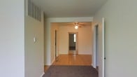 This single-floor residence offers a straightforward and functional layout designed for comfortable everyday living. The interior opens into a welcoming living area adorned with warm hardwood flooring that extends throughout the main spaces, lending the home a cohesive and timeless aesthetic. A ceiling fan is centrally positioned, providing both practical ventilation and a subtle design element to the room.

The layout flows naturally from one area to the next, with wide doorways connecting the main living space to adjoining rooms, offering an open and accessible feel throughout. A carpeted entry or transitional zone adds a layer of texture and warmth underfoot, differentiating spaces within the home.

Neutral-toned walls throughout the residence create a clean, versatile backdrop that allows for a variety of interior design choices. Visible HVAC venting integrated into the upper wall indicates a climate-controlled environment for year-round comfort. A doorway at the far end of the main living area reveals a glimpse of a room with natural light filtering through a window, hinting at bright, airy spaces deeper within the home. An additional door, likely leading to a closet or utility space, adds to the practical storage offerings of this residence. The overall ambiance is calm, clean, and move-in ready.