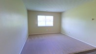 This single-floor space offers a straightforward and functional layout with a clean, move-in-ready aesthetic. The main room is a generously sized, open area finished with wall-to-wall carpeting in a neutral tone, providing a comfortable and quiet underfoot experience. The walls are painted in a soft, warm beige that lends the space a calm and inviting atmosphere. A centrally positioned window on the far wall allows natural light to fill the room, brightening the interior and offering a view of the surrounding outdoor environment. White baseboards provide a crisp contrast against the light-colored walls and carpet, contributing to the room's tidy, well-maintained appearance. Electrical outlets are conveniently placed along the walls, offering flexibility for furniture arrangement and everyday use. The ceiling is smooth and white, enhancing the sense of openness within the room. Overall, the space presents a blank canvas with a neutral palette and practical finishes, making it highly adaptable to a variety of uses and personal styles. Its simplicity and clean condition make it an accessible and versatile option for prospective occupants.