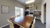 This single-floor space functions as a versatile meeting and work room, thoughtfully designed to accommodate both collaborative gatherings and individual tasks. The central feature is a substantial wood-topped conference table surrounded by white ladder-back chairs, providing ample seating for group meetings or shared activities. Patterned carpet in a neutral gray-blue tone covers the floor throughout, adding texture and a professional feel to the room.

Built-in shelving units along one wall offer organized storage and display space, housing books, decorative accents, and various items. Adjacent to the shelving, a dedicated workstation area includes a desktop computer and printer, making the room suitable for administrative or business use. A large window fitted with vertical blinds allows natural light to filter into the space while offering view to the outdoors.

A vibrant, colorful piece of wall art depicting a tree-lined pathway serves as a focal point and adds a welcoming, lively touch to the otherwise neutral interior. The room connects directly to a carpeted hallway corridor through glass-paneled double doors, suggesting this space is part of a larger facility. Overhead fluorescent lighting ensures the room is well-lit for functional use throughout the day. Overall, the space strikes a balance between practical utility and a comfortable, inviting atmosphere.