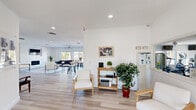 This single-floor community clubhouse offers a generously proportioned open-plan layout designed for both relaxation and social gatherings. Light-toned luxury vinyl plank flooring runs throughout, complementing the crisp white walls and recessed lighting that create a clean, welcoming atmosphere.

The main living area serves as the focal point of the space, anchored by a striking floor-to-ceiling white marble-tile feature wall housing a built-in fireplace and wall-mounted television. Comfortable seating arrangements and a ceiling fan overhead add to the inviting ambiance. Large sliding windows and French doors flood this area with natural light while framing panoramic views of the outdoor swimming pool and surrounding palm-lined grounds.

Adjacent to the lounge, a versatile gathering zone offers additional seating with bar-height tables and a built-in counter with bar stools — ideal for casual meetings or events. Louvered partition doors provide flexible separation when needed.

The fully equipped kitchen features crisp white shaker cabinetry, a classic subway tile backsplash, stainless steel appliances including a French-door refrigerator and microwave, and ample counter space. A welcoming entry foyer with French doors opening to the outdoors completes the layout, enhancing the seamless indoor-outdoor flow throughout this well-appointed community space.