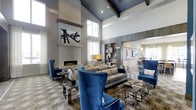 This single-floor space makes an immediate impression with its soaring vaulted ceilings accented by dark exposed beams and a rich navy blue upper ceiling, creating a dramatic sense of height and grandeur. The focal point of the main living area is a floor-to-ceiling stacked stone fireplace with a sleek linear firebox, flanked by tall windows that flood the space with natural light and sheer patterned drapery that softens the brightness.

The interior design balances bold and neutral tones seamlessly. Royal blue velvet accent chairs with nailhead trim command attention alongside a taupe upholstered sofa, all anchored by a large patterned area rug in warm earth tones. Mirrored and metallic accent tables add a polished, contemporary edge throughout. A built-in shelving unit with decorative objects provides both function and visual interest along one wall.

An open passage leads to an adjacent room, suggesting a fluid, connected layout ideal for entertaining. The overall ambiance is refined and modern, combining architectural drama with curated furnishings to achieve a space that feels both expansive and inviting.