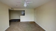 This single-floor residence offers a straightforward and functional layout designed to maximize usable living space. The main living area is finished with wall-to-wall carpet in a neutral beige tone, creating a warm and understated atmosphere throughout. A ceiling fan with an integrated light fixture is centrally mounted, providing both air circulation and ambient lighting to the room.

The living space flows seamlessly into the kitchen area, which is separated by a raised breakfast bar or pass-through counter. This semi-open layout allows for easy interaction between the two spaces while maintaining a sense of definition. The kitchen is equipped with standard appliances, including what appears to be an oven and range, and features dark cabinetry that contrasts with the lighter walls and surfaces.

The entryway to the unit is visible near the kitchen, with a standard interior door finished in white. The overall color palette of the space is clean and minimal, with light-colored walls throughout that contribute to a bright and airy feel. The transition from carpeted living areas to hard flooring near the kitchen entry provides a practical delineation between spaces. This unit offers a simple, no-frills layout suitable for comfortable everyday living.