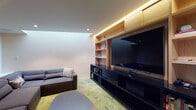 Located at 201 NE 2nd Ave in Portland, Oregon, this single-floor media lounge is thoughtfully designed for comfort and entertainment. The room's most striking architectural feature is a large angled skylight set into the ceiling, which floods the otherwise intimate space with diffused natural light, creating a bright and airy atmosphere despite the room's cozy scale.

Dominating one full wall is a custom-built entertainment unit crafted from warm natural wood, featuring open shelving for books and décor, upper cabinet storage with integrated LED cove lighting, and a sleek black lower media console that houses the large flat-screen television. The contrast between the light wood millwork and the dark media console gives the wall a polished, contemporary feel.

The seating area is generously appointed with a large, low-profile modular sectional sofa upholstered in charcoal fabric, capable of accommodating multiple guests with ease. An oval coffee table in dark wood anchors the center of the room. Underfoot, a textured green-toned carpet adds warmth and a subtle pop of color to the neutral palette of white walls and recessed ceiling lighting. A discreet entry door completes the layout, making this a well-appointed, self-contained media and lounge space.