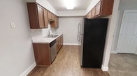 This single-floor residence offers a functional and thoughtfully designed layout. The kitchen stands out as a central feature, showcasing warm medium-toned wood cabinetry that lines both the upper and lower walls, providing generous storage throughout. Light-toned countertops with a subtle pattern complement the cabinetry and offer ample workspace for meal preparation. The kitchen is equipped with stainless steel appliances, including a dishwasher and a full-sized refrigerator, adding a modern touch to the space. A stainless steel sink with a single-handle faucet is neatly integrated into the counter, positioned beneath the upper cabinets for convenience. The flooring throughout features durable hardwood-style planks in a warm, natural tone that ties the space together cohesively. Neutral gray walls provide a clean, versatile backdrop that complements the wood tones and bright finishes. The textured ceiling adds a classic residential character to the space. Overall, the kitchen presents a practical yet polished environment, balancing traditional wood detailing with contemporary appliances for everyday comfort and functionality.