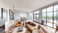 Located at 212 Durham Ave in Metuchen, NJ, this single-floor space seamlessly integrates a living room and dining area into one cohesive, design-forward environment. The living area is anchored by a sleek, wall-mounted fireplace with a dark surround, above which a flat-screen TV is mounted. Curated artwork, including a large abstract canvas with layered horizontal strokes, adds artistic character to the crisp white walls. A bold rust-and-white patterned area rug grounds the lounge seating, which includes a curved sectional and organic sculptural accent chairs. Floor-to-ceiling black-framed glass doors and windows flood the space with natural light and open directly to an outdoor terrace with string lighting and exterior seating. The dining area features a striking geometric black-and-white tile floor, an oval white dining table surrounded by sculptural wood-and-leather chairs, and a dramatic tray ceiling with a cascading multi-cone brass pendant light. A compact kitchen with dark tile backsplash, stainless steel appliances, and white cabinetry is integrated into the dining zone. The space maintains a modern, curated ambiance throughout, with thoughtful lighting and an abundance of greenery views from the surrounding windows.