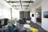 This contemporary communal lounge is thoughtfully designed to offer both comfort and style across a single open floor. The space is anchored by a dramatic floor-to-ceiling stone-tile fireplace wall, which serves as a striking focal point and lends a sense of warmth and architectural sophistication to the room. A coffered ceiling with metallic accents and pendant lighting adds visual depth and elegance overhead.

Natural light floods the interior through expansive floor-to-ceiling windows that wrap along multiple sides of the space, offering panoramic views of the surrounding cityscape. The windows are complemented by a built-in bar-height counter and seating area, creating an ideal perch for taking in the scenery.

The main seating area features a generously sized sectional sofa in charcoal gray, enlivened by bold teal and yellow accent pillows, paired with modern rounded ottomans and minimalist side tables. A chevron-patterned carpet grounds the lounge area, adding texture and visual interest underfoot.

Beyond the main lounge, the space opens to a secondary area with wall-mounted displays and access to what appears to be a kitchenette or bar space. Exposed concrete columns punctuate the room, reinforcing the building's modern structural character. The overall ambiance is bright, polished, and inviting, making it a well-appointed gathering space suited for both relaxation and social interaction.