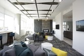 This contemporary communal lounge is thoughtfully designed to offer both comfort and style across a single open floor. The space is anchored by a dramatic floor-to-ceiling stone-tile fireplace wall, which serves as a striking focal point and lends a sense of warmth and architectural sophistication to the room. A coffered ceiling with metallic accents and pendant lighting adds visual depth and elegance overhead.

Natural light floods the interior through expansive floor-to-ceiling windows that wrap along multiple sides of the space, offering panoramic views of the surrounding cityscape. The windows are complemented by a built-in bar-height counter and seating area, creating an ideal perch for taking in the scenery.

The main seating area features a generously sized sectional sofa in charcoal gray, enlivened by bold teal and yellow accent pillows, paired with modern rounded ottomans and minimalist side tables. A chevron-patterned carpet grounds the lounge area, adding texture and visual interest underfoot.

Beyond the main lounge, the space opens to a secondary area with wall-mounted displays and access to what appears to be a kitchenette or bar space. Exposed concrete columns punctuate the room, reinforcing the building's modern structural character. The overall ambiance is bright, polished, and inviting, making it a well-appointed gathering space suited for both relaxation and social interaction.