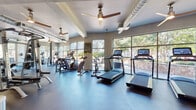 This fitness center offers a comprehensive workout environment designed to accommodate a wide range of exercise needs. The space is organized into distinct functional zones, seamlessly flowing from one area to the next across a single open floor.

The cardio section is lined with modern treadmills, elliptical trainers, and stationary bikes, all positioned to take advantage of the large floor-to-ceiling windows that flood the area with natural light and provide views of a lush outdoor courtyard and patio space. The expansive glazing creates an open, airy atmosphere that makes workouts feel less confined.

The strength training area features a full complement of free weights including dumbbells and barbells, weight plates, adjustable benches, and a multi-station cable and rack system. Full-length mirrors line the walls of this section, enhancing the sense of space while allowing users to monitor their form.

Throughout the facility, ceiling fans with distinctive gold and brushed metal finishes are mounted to the structured ceiling, which showcases exposed ductwork as an industrial design element. The durable rubber flooring transitions from blue-toned in the cardio zone to dark charcoal in the strength area, providing both safety and a polished aesthetic. Glass-paneled entry doors and neutral gray walls complete the clean, modern design of this well-appointed fitness facility.