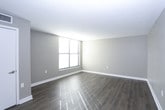 This single-floor unit presents a functional and well-organized layout, as revealed by its overhead floor plan view. The living area is finished with light wood-tone flooring and neutral beige walls, creating a clean, understated aesthetic. Natural light filters in through window blinds, adding brightness to the open space. White baseboards throughout provide a crisp contrast to the warm-toned flooring.

The kitchen is efficiently arranged with rich, dark cherry wood cabinetry offering ample upper and lower storage. It is equipped with black appliances, including a refrigerator, electric range, built-in microwave, and dishwasher. Light-toned countertops complement the cabinetry, while a neutral tile backsplash adds texture to the cooking area. Wood-tone flooring continues seamlessly from the living spaces into the kitchen.

The bedroom features the same neutral wall palette, with carpet flooring for added comfort. A window with horizontal blinds allows for natural light and ventilation. The bathroom includes a soaking tub fitted with large-format, earth-toned stone-look tiles covering the surrounding walls, lending a grounded, durable finish to the space. Overall, the unit maintains a cohesive, move-in-ready aesthetic throughout.