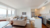 This thoughtfully designed single-floor residence showcases a bright, open-concept layout with clean white walls, smooth ceilings, and rich dark hardwood flooring that unifies the living spaces. The living area is generously proportioned, bathed in natural light streaming through large floor-to-ceiling windows that frame sweeping city skyline views. A comfortable seating arrangement anchors the space, complemented by a dedicated dining area with a round wood table and seating for four.

The kitchen is a standout feature, offering stainless steel appliances including a refrigerator, gas range, and built-in microwave, set against a distinctive geometric hexagonal tile backsplash. Light wood cabinetry lines the walls, providing ample storage, while a central island with a butcher-block countertop and white bar stools serves as a casual dining and prep space.

The bedroom continues the home's contemporary aesthetic, with large windows offering expansive city views and dark wood flooring underfoot. The bathroom features a full soaking tub and shower combination, accented by hexagonal wall tiles that echo the kitchen's design language, creating a cohesive style throughout. The overall ambiance is modern, airy, and light-filled, with a neutral palette that highlights the urban panorama beyond.