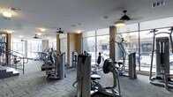 This fitness center offers a comprehensive workout environment outfitted with a wide range of exercise equipment. The space features a strong selection of cardio machines, including ellipticals and steppers, as well as strength training equipment positioned throughout the room. Large, floor-to-ceiling windows line the perimeter, flooding the space with natural light while providing open views of the surrounding streetscape, creating an energizing atmosphere for workouts. The windows also lend a sense of openness and connectivity to the urban environment outside. Ceiling fans are installed throughout, supporting air circulation and comfort during use. The flooring consists of a patterned carpet, providing a functional and durable surface suited to fitness activity. Warm-toned accent walls contrast with the bright white ceiling, contributing to an inviting yet focused atmosphere. Recessed and flush-mount lighting supplements the natural light, ensuring the space remains well-lit at all hours. The layout is thoughtfully organized, with equipment arranged to allow ample circulation space between machines. Overall, the fitness center is designed to support a productive and comfortable exercise experience.