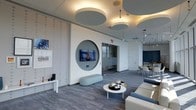 This single-floor space showcases a thoughtfully designed interior with a contemporary aesthetic defined by a cohesive blue-grey color palette. The lounge area serves as a central focal point, featuring a striking circular wall alcove that frames a wall-mounted flat-screen TV, creating a sculptural architectural detail that draws the eye immediately upon entry. Complementing this design element, the ceiling is adorned with large circular acoustic panels finished in soft grey and white tones, providing both visual interest and sound management throughout the space.

The flooring consists of wood-look planks in a cool grey tone, lending warmth while maintaining the modern character of the interior. Seating arrangements include a curved light-blue bench, a round white coffee table with decorative accents, and plush upholstered sofas in muted blue-grey hues. A textured accent wall with a subtle dot pattern adds depth to the lounge area, while curated artwork and decorative pieces contribute to the polished atmosphere.

Beyond the lounge, a glimpse into an adjacent dining area reveals round tables and chairs alongside a bold navy blue accent wall, suggesting a versatile, multi-functional layout designed for both relaxation and social gathering.