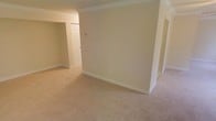 This single-floor space offers a straightforward and functional layout with a warm, neutral aesthetic. The interior is finished with soft beige wall tones and plush wall-to-wall carpeting that adds comfort underfoot while contributing to a cohesive, understated color palette throughout.

Crown molding runs along the ceiling, providing a classic architectural detail that adds a subtle sense of refinement to the space. The arched doorway visible in the image introduces a traditional design element, lending gentle visual interest and helping to define the transition between areas.

The layout includes multiple connected rooms, with doorways leading to additional spaces such as what appears to be a closet or storage area with a paneled door. White baseboards frame the floors cleanly, complementing the bright trim work found throughout. Electrical outlets are positioned at standard heights along the walls, and a thermostat or control panel is mounted conveniently within reach.

Overall, the space presents a blank canvas with a clean, move-in-ready condition. Its neutral finishes, classic detailing, and practical layout make it well-suited for a variety of furnishing styles and personal customizations.