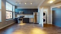 Located at 23 Central in Lynn, Massachusetts, this modern single-floor unit offers a thoughtfully designed layout with contemporary finishes and practical amenities. The kitchen is a standout feature, outfitted with dark wood cabinetry, stainless steel appliances including a range, microwave, dishwasher, and refrigerator, as well as light quartz countertops and a classic subway tile backsplash. Track lighting illuminates the space, highlighting the warm wood-tone flooring that flows throughout. A dedicated in-unit laundry closet with a stacked washer and dryer adds everyday convenience directly off the kitchen area. The living area is bright and open, benefiting from large windows that frame views of the surrounding urban streetscape, allowing natural light to fill the room and create an airy atmosphere. The bathroom is clean and functional, featuring light tile flooring, a white vanity with an undermount sink, a mirror with overhead lighting, and a combined tub and shower. The overall design balances modern aesthetics with practical functionality, making this a well-appointed urban residence in the heart of Lynn's central district.