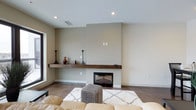 This single-floor, 2-bedroom, 2-bathroom residence offers a thoughtfully designed living space defined by a contemporary aesthetic. The living room serves as the heart of the unit, anchored by a built-in fireplace framed by a substantial dark wood floating mantel shelf that provides both visual warmth and a display surface. Neutral beige and greige wall tones create a calm, cohesive backdrop throughout. Recessed lighting in the smooth ceiling provides clean, even illumination, complementing the natural light that filters in through a large window visible along the left wall. The flooring features polished concrete-style surfaces in a cool, dark tone, lending an urban, modern character to the space. The open layout of the living area accommodates comfortable furnishings, including a plush area rug that softens the hard flooring. The overall ambiance is understated and refined, balancing minimalist design with welcoming elements. The combination of the fireplace, warm wood accents, and neutral palette gives the space a grounded, livable quality suited to both relaxing and entertaining.