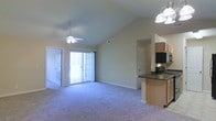 This single-story residence showcases an open-concept layout that seamlessly connects the living area and kitchen. The living space is defined by striking vaulted ceilings that rise dramatically overhead, enhancing the sense of volume and airiness throughout the room. A ceiling fan is centrally mounted, providing both practical ventilation and a finishing architectural detail. Neutral beige walls and wall-to-wall carpeting in a soft gray-blue tone create a calm, cohesive atmosphere throughout the main living area.

Natural light flows generously into the space through a large sliding glass door, which also provides direct access to an outdoor area. A traditional paneled door on the opposite side of the room offers an additional entry point.

The adjacent kitchen is partially open to the living area, separated by a raised breakfast bar with a dark countertop that provides both a visual boundary and a casual dining surface. The kitchen features warm wood-toned cabinetry, dark countertops, and includes a range and overhead microwave. A pendant light fixture with a multi-bulb design hangs above, illuminating the workspace effectively. Overall, the space presents a functional and comfortable single-floor layout.