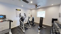 This single-floor fitness room offers a practical and well-organized space for exercise. The room is finished with light gray walls and dark rubber flooring, providing a clean, functional aesthetic suited to a workout environment. Recessed lighting is complemented by two ceiling fans, ensuring the space remains well-lit and comfortable during use. A window with blinds allows natural light to filter in while maintaining privacy.

The room is equipped with a selection of cardio and strength training equipment, including an elliptical machine, a recumbent stationary bike, and an upright exercise bike, all positioned to allow ease of movement between stations. A weight bench adds versatility for strength training. A posted "Fitness Area Rules" sign near the entrance provides guidance for users. A door at the rear of the room leads to an additional area, potentially a restroom or storage space, adding further functionality. The overall layout is efficient, making good use of the available space while keeping the environment tidy and accessible.
