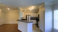 This single-floor space showcases an open-concept layout that seamlessly connects the kitchen and living areas. The kitchen is a standout feature, outfitted with crisp white shaker-style cabinetry that extends to the upper walls, providing generous storage throughout. Dark countertops offer a striking contrast against the white cabinetry, and a peninsula with matching dark countertop extends into the space, creating a natural division between the kitchen and the adjoining living area while also serving as a functional breakfast bar or prep surface. Stainless steel appliances, including a refrigerator, range, and built-in microwave, lend a modern, cohesive aesthetic. The flooring transitions from light tile in the kitchen to warm hardwood in the living area, subtly defining each zone. Crown molding runs along the ceiling throughout, adding a refined architectural detail. A decorative column near the peninsula adds structural character to the open floor plan. Track lighting above the kitchen provides focused illumination, complementing the overall bright and functional ambiance of the space.