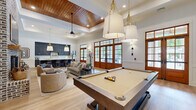 Located at 5171 Bowman Rd in Macon, Georgia, this single-floor entertainment space is thoughtfully designed to blend elegance with functionality. The open-concept layout accommodates multiple activity zones seamlessly, highlighted by rich light oak hardwood flooring that runs throughout. A dramatic floor-to-ceiling whitewashed brick fireplace serves as the room's focal point, anchoring a comfortable lounge area furnished with neutral sofas and curved accent chairs. Adjacent to the lounge, a full-size pool table takes center stage beneath a cluster of oversized drum pendant lights with brass accents, adding both warmth and sophistication. The ceiling features a striking combination of a warm wood-paneled tray inset and crisp white crown molding, contributing to a sense of elevated height and architectural refinement. Stately wood-framed French doors line the walls, inviting natural light and providing views of the lush outdoor surroundings. The wet bar and kitchenette area is finished with deep navy shaker-style cabinetry, white quartz countertops, a large island with rattan barstools, and gold pendant lighting — creating a polished, high-end aesthetic. Wainscoting details along the lower walls add a classic Southern charm that ties the entire space together cohesively.