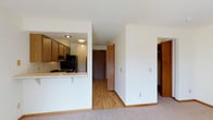 This single-floor residential unit offers a practical and well-organized layout designed for comfortable everyday living. The space features a semi-open kitchen defined by a pass-through counter with a breakfast bar overhang, providing a natural connection between the kitchen and the living area. The kitchen is equipped with wood-toned cabinetry, a gas range, and a full-size refrigerator, with recessed and flush-mount lighting providing consistent illumination throughout. Light hardwood flooring runs through the entry corridor and kitchen area, while plush carpeting covers the living space, creating a warm and welcoming transition between zones. Crisp white walls throughout the unit enhance the sense of openness and keep the interior feeling bright. A central hallway leads toward the main entry door, which features warm wood paneling that complements the kitchen cabinetry. To the right, a built-in closet with wood shelving offers generous storage. The clean, neutral palette and straightforward design make the space highly adaptable to a variety of furnishing styles. Overall, the unit presents a tidy, functional environment with cohesive warm-toned wood accents tying the interior together.