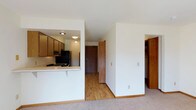 This single-floor residential unit offers a practical and well-organized layout designed for comfortable everyday living. The space features a semi-open kitchen defined by a pass-through counter with a breakfast bar overhang, providing a natural connection between the kitchen and the living area. The kitchen is equipped with wood-toned cabinetry, a gas range, and a full-size refrigerator, with recessed and flush-mount lighting providing consistent illumination throughout. Light hardwood flooring runs through the entry corridor and kitchen area, while plush carpeting covers the living space, creating a warm and welcoming transition between zones. Crisp white walls throughout the unit enhance the sense of openness and keep the interior feeling bright. A central hallway leads toward the main entry door, which features warm wood paneling that complements the kitchen cabinetry. To the right, a built-in closet with wood shelving offers generous storage. The clean, neutral palette and straightforward design make the space highly adaptable to a variety of furnishing styles. Overall, the unit presents a tidy, functional environment with cohesive warm-toned wood accents tying the interior together.