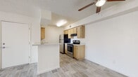 This single-story residence is designed with a practical, open layout that connects the main living and kitchen areas seamlessly. Light-toned wood-look flooring extends consistently throughout the unit, contributing to a clean, cohesive aesthetic.

The kitchen is a functional galley-style space equipped with warm oak-toned cabinetry, stainless steel appliances including an electric range and refrigerator, a built-in dishwasher, and a double-basin sink. A peninsula with a light stone-look countertop separates the kitchen from the living area, providing additional workspace and a casual dining option.

The living area is bright and open, featuring a textured ceiling, a ceiling fan, and a sliding glass door with vertical blinds that allows natural light and provides access to the exterior. A second room, also with a ceiling fan and textured ceiling, offers versatile use as a bedroom or additional living space.

The bathroom includes a wide vanity with an oak cabinet base, a speckled countertop, and a large mirror. A dedicated laundry closet houses a stacked washer and dryer unit with built-in shelving nearby for added storage convenience.