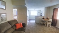 This single-floor space is designed with a practical, open layout that connects the main living areas seamlessly. The living room is carpeted and centers around a comfortable seating arrangement, with a floor vent providing climate control and neutral beige walls creating a warm, understated atmosphere. A textured ceiling runs throughout, adding a classic residential character to the space.

From the living area, sightlines extend directly into the dining nook, which features a compact round table and chairs positioned near the pass-through kitchen counter. The kitchen is partially visible from the main living space via an open bar-style counter, and is equipped with white cabinetry and an overhead range hood. A window above the kitchen area allows natural light to filter into the space, brightening the interior.

A hallway visible from the living room leads to the bedroom, which features a bed with light-toned bedding and curtained windows. The transition areas display hard flooring, contrasting with the carpeted living and sleeping zones. Thermostats mounted along the hallway wall indicate individual climate control access throughout the unit.