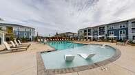 Located at 3100 Ascend Loop in Leland, North Carolina, this outdoor amenity area offers a resort-inspired pool experience designed for relaxation and recreation. The centerpiece is a generously sized swimming pool with crystal-clear blue water, complemented by a shallow wading area featuring built-in seating — ideal for leisurely lounging in the water. The expansive pool deck is finished in a warm, neutral concrete surface and is lined with rows of sling-style chaise lounges and side tables, providing ample space for sunbathing and outdoor socializing. Several shade umbrellas are positioned throughout the deck for added comfort. Tropical palm trees and carefully curated landscaping with colorful shrubs and ornamental plantings add a vibrant, coastal ambiance to the space. The area is enclosed by a black iron perimeter fence, ensuring both safety and a clean aesthetic. Surrounding multi-story residential buildings with classic coastal architecture — featuring gray and blue siding, pitched roofs, and large windows — frame the pool area on multiple sides, creating a cohesive community feel. The overall atmosphere is bright, open, and inviting, making this outdoor space a standout amenity within the community.
