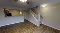 This two-story home offers a well-organized layout designed for comfortable living. The main level features an open-concept living area finished with rich, dark hardwood-style flooring that runs throughout, adding warmth and visual continuity to the space. Neutral gray-toned walls provide a clean, modern backdrop that complements the flooring and trim work.

A ceiling fan with a light fixture serves the living area, offering both airflow and ambient lighting. The space flows naturally toward a kitchen area, partially visible through a pass-through counter topped with a light-colored stone or laminate surface. The kitchen includes a sink positioned near a window, allowing natural light into the area. A doorway beyond the kitchen suggests additional rooms or utility spaces on this level.

The staircase, centrally positioned, features white-painted balusters and a warm wood handrail, leading to the second floor. Crown molding and clean baseboards throughout the main level reflect attention to detail in the finishwork. Overall, the space presents a functional and inviting interior with a cohesive design aesthetic suited for everyday living.