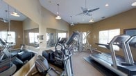 This fitness center offers a comprehensive workout environment designed to accommodate a wide range of exercise needs. The space is thoughtfully laid out across a single floor, with a generous open layout that provides ample room to move between equipment without feeling cramped. The flooring is carpeted throughout, lending a softer, more comfortable underfoot experience compared to typical gym settings. A diverse selection of cardio machines is prominently featured, including ellipticals, a stair climber, and a treadmill, catering to various aerobic training preferences. Strength training is well represented with a multi-station cable and weight machine, along with additional resistance equipment. Large mirrors span one full wall, visually expanding the space and allowing users to monitor their form during workouts. Natural light filters in through multiple windows, creating a bright and energizing atmosphere. Overhead, ceiling fans work in tandem with pendant lighting fixtures to maintain a comfortable, well-lit environment. The warm, neutral-toned walls contribute to an inviting ambiance. The overall design balances functionality with a clean, organized aesthetic, making this fitness center a practical and welcoming space for daily use.