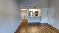 This single-floor space showcases a clean, modern design with an open-concept layout that connects the living area to the kitchen seamlessly. The flooring throughout is warm-toned hardwood, adding a cohesive and polished look to the interior. Notably tall ceilings lend an airy, spacious feel to the unit.

The kitchen is outfitted with white cabinetry, a granite-topped peninsula that doubles as a breakfast bar, and a full suite of stainless steel appliances, including a refrigerator, microwave, and range. The peninsula provides additional prep space while creating a natural divide between the kitchen and the main living area.

Accessible directly from the main living space is a dedicated laundry area, complete with a full-size washer and dryer — a practical and convenient feature. The neutral gray walls throughout complement the light-colored cabinetry and bright finishes, creating a fresh and modern ambiance. HVAC vents are integrated into the ceiling, ensuring efficient climate control. The overall aesthetic is functional yet contemporary, offering a well-organized and comfortable living environment.