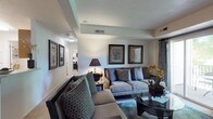 This single-floor residence offers a thoughtfully designed living space that balances comfort and sophistication. The living room serves as the centerpiece of the home, showcasing a distinctive tray ceiling that adds architectural depth and visual interest to the room. Neutral wall tones provide a versatile backdrop, complemented by warm, layered lighting from ceiling fixtures and accent lamps that create an inviting atmosphere. The space is furnished with a plush, light-toned sofa accented by teal and patterned throw pillows, alongside a coordinating loveseat, establishing a cohesive and stylish seating arrangement. A sleek glass-top coffee table anchors the sitting area, while a decorative arrangement of fresh flowers adds a vibrant, natural touch. The walls are adorned with framed botanical artwork, enhancing the refined aesthetic of the room. Elegant drapery frames the windows, providing privacy while adding to the polished décor. A hallway visible from the living area hints at additional rooms beyond, suggesting a well-organized and functional floor plan. Overall, this space presents a harmonious blend of classic design elements and contemporary comforts, making it a welcoming and visually appealing environment.