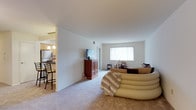 This single-floor residence offers an open and functional layout that connects the main living area to the kitchen. The living room is generously sized, featuring neutral wall-to-wall carpeting and white walls that create a bright, clean atmosphere. A large window with vertical blinds allows ample natural light to fill the space, enhancing its airy feel. The room comfortably accommodates a curved sectional sofa and an entertainment unit housing a flat-screen television, reflecting its practical design. Adjacent to the living room, the kitchen is partially separated by a half-wall partition, maintaining an open connection between the two areas. The kitchen features light wood cabinetry, a dark countertop breakfast bar with bar stools, and pendant lighting overhead, adding a warm contrast to the neutral palette. The transition between the carpeted living space and the hard-floored kitchen is clearly defined, contributing to the overall organized flow of the layout. Together, the spaces offer a cohesive and comfortable environment with a straightforward, livable design suited for everyday use.