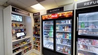 This single-floor micro market is designed to provide convenient, self-service retail within a compact and efficiently organized space. The interior features a drop-tile ceiling with recessed panel lighting that keeps the area bright and well-illuminated throughout. The flooring consists of light-toned tile, contributing to a clean and functional aesthetic.

The retail area is organized into clearly labeled sections, including dedicated "Snack," "Quench," and "Refuel" zones. The snack section offers shelving stocked with a wide variety of packaged chips, candy, and other grab-and-go items. The "Quench" zone is anchored by a large, double-door refrigerated unit prominently branded with Coca-Cola signage, stocked with an extensive selection of cold beverages including sodas, juices, and water. Adjacent to it, the "Refuel" refrigerator provides chilled food items such as sandwiches and other ready-to-eat options.

A self-checkout station equipped with a digital screen and point-of-sale kiosk provides a seamless, cashier-free purchasing experience, supported by Three Square Market's platform. Informational signage near the checkout area guides users on setting up their accounts. The space also includes a secured interior door with a wire-reinforced window, likely leading to a back room or restricted area. The overall layout is practical, accessible, and optimized for quick, self-directed shopping.
