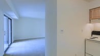 This single-floor space presents a clean, minimalist aesthetic characterized by crisp white walls and soft, plush carpeting throughout. The interior is bright and airy, with an open layout that allows for flexible use of the available square footage. The design is simple and uncluttered, providing a neutral canvas well-suited for a variety of furnishing styles and configurations. A wide doorway or passageway connects adjoining areas, enhancing the sense of flow and openness between rooms. Standard electrical outlets are conveniently positioned along the walls, reflecting practical, move-in-ready functionality. The consistent light color palette across walls and flooring amplifies the natural light within the space, contributing to a calm and welcoming atmosphere. The overall condition of the interior appears well-maintained, with smooth wall finishes and evenly laid carpeting. The straightforward, unadorned design makes this space highly adaptable, whether for residential or light professional use. The single-level layout ensures easy accessibility throughout, with no stairs to navigate between areas.