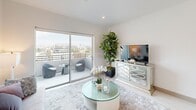 This contemporary urban residence is thoughtfully designed with clean lines, neutral tones, and high-end finishes that create a bright, cohesive aesthetic throughout. The kitchen is a standout feature, offering crisp white upper cabinetry, warm wood-toned lower cabinets, stainless steel appliances, a decorative tile backsplash, and a mobile island with a white quartz surface — all illuminated by a stylish pendant light and recessed lighting. The living area is airy and inviting, with large sliding glass doors that open onto a private balcony, framing sweeping city skyline views and flooding the space with natural light. The balcony provides an ideal outdoor retreat with ample room for seating. The primary bedroom features a walk-in closet and large windows offering additional urban views, while the secondary bedroom also captures city vistas through a generous window. Both bedrooms are finished with clean, neutral walls and recessed lighting. The bathroom is appointed with wood-look tile flooring, a floating vanity with a white countertop, an LED-backlit mirror, and a tiled shower and tub surround. Throughout the space, light tile flooring and white walls enhance the sense of openness, creating a polished and modern living environment with an urban edge.
