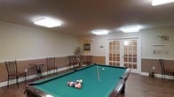 This single-floor recreational space is thoughtfully designed to provide a welcoming environment for leisure and social activity. The centerpiece of the room is a full-size billiards table with a classic green felt surface, positioned prominently beneath a series of flush-mounted overhead light fixtures that provide bright, even illumination throughout the space. The room features a two-tone wall treatment, with a warm neutral upper half and a deeper wainscoting-style lower section, adding a touch of elegance to the casual setting. Hardwood-style flooring runs throughout, complementing the warm color palette of the walls and ceiling, which features a subtle textured finish. Along the perimeter of the room, wrought iron chairs offer casual seating for spectators or those waiting their turn at the table, while a decorative floral arrangement adds a welcoming touch. A standout architectural feature is the set of classic French doors with divided glass panes, leading to an adjoining area and allowing light to flow between spaces. Informational and instructional posters on the walls contribute to the room's functional character. Overall, the space balances practicality with comfort, making it an ideal gathering spot for recreation and socializing.