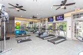 This fitness center is a thoughtfully designed, single-floor facility built to support a comprehensive workout experience. The space is generously proportioned, offering room for both cardiovascular and strength training activities without feeling cramped. A row of True-brand treadmills and cardio machines lines one side, while a variety of weight and resistance training equipment occupies the central and opposite areas. Large windows along one wall flood the room with natural light, creating a bright and energizing atmosphere, while multiple glass-paneled doors provide easy access and enhance the open feel. Ceiling fans are evenly distributed throughout, ensuring consistent airflow and comfort during workouts. Wall-mounted flat-screen TVs are positioned for visibility from multiple workout stations, adding an entertainment element to training sessions. A floor-to-ceiling mirror spans one wall, visually expanding the space and allowing users to monitor their form. Recessed lighting supplements the natural light, keeping the facility well-illuminated at all hours. The flooring is durable and fitness-appropriate, designed to withstand heavy equipment and high foot traffic. Overall, the space presents a clean, functional, and welcoming environment suitable for a wide range of fitness routines.