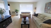 This single-floor space offers a welcoming and functional layout centered around a generously sized living room. The room is finished with soft, neutral-toned carpeting that adds warmth underfoot, while crisp white walls and a smooth ceiling create a clean, open atmosphere throughout.

The living room is a standout feature, anchored by a large sliding glass door dressed with vertical blinds that allow abundant natural light to pour into the space. The door opens to an outdoor area, providing both a visual connection to the outside and easy access to fresh air. Two comfortable sofas are arranged around a sleek, dark coffee table, creating an inviting seating arrangement ideal for relaxing or entertaining.

Decorative touches — including framed wall art, a table lamp, and potted plants — add character and a lived-in warmth to the space. A wall-mounted air conditioning unit ensures year-round comfort. The overall ambiance is light, airy, and practical, with a neutral color palette that makes the space feel open and easy to personalize. The layout flows naturally, making efficient use of the available space while maintaining a comfortable and approachable feel throughout.