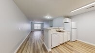 This single-floor residence offers a well-organized layout accommodating three bedrooms, two bathrooms, a combined kitchen and living area, a hallway, and a closet. The open-concept kitchen and living room serve as the central hub of the home. The kitchen features white cabinetry, a marble-look countertop peninsula that doubles as a breakfast bar, white appliances including a refrigerator, dishwasher, and microwave, and a double-basin sink. Light wood-style vinyl plank flooring runs throughout the main living areas, contributing to a clean, cohesive aesthetic. The spacious living room receives natural light through a large window, enhancing the airy feel of the soft gray-blue painted walls.

The three bedrooms are finished with neutral carpet and each includes a window with blinds, providing natural light. The primary bedroom offers a generous footprint among the three. Both bathrooms are finished with wood-style flooring and feature white fixtures, including a tub-shower combination. One bathroom includes a white vanity with under-sink storage and a mirror with overhead lighting. The overall ambiance is clean, bright, and move-in ready with a consistent neutral tone throughout.