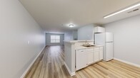 This single-floor residence offers a well-organized layout accommodating three bedrooms, two bathrooms, a combined kitchen and living area, a hallway, and a closet. The open-concept kitchen and living room serve as the central hub of the home. The kitchen features white cabinetry, a marble-look countertop peninsula that doubles as a breakfast bar, white appliances including a refrigerator, dishwasher, and microwave, and a double-basin sink. Light wood-style vinyl plank flooring runs throughout the main living areas, contributing to a clean, cohesive aesthetic. The spacious living room receives natural light through a large window, enhancing the airy feel of the soft gray-blue painted walls.

The three bedrooms are finished with neutral carpet and each includes a window with blinds, providing natural light. The primary bedroom offers a generous footprint among the three. Both bathrooms are finished with wood-style flooring and feature white fixtures, including a tub-shower combination. One bathroom includes a white vanity with under-sink storage and a mirror with overhead lighting. The overall ambiance is clean, bright, and move-in ready with a consistent neutral tone throughout.