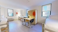 Located at 525 Lexington Avenue in Midtown Manhattan, this efficiently designed room offers a thoughtfully organized living environment on a single floor. The main living area accommodates multiple beds with built-in storage drawers underneath, maximizing the use of available space. A shared work surface with rolling chairs provides a dedicated study zone, while a tall wardrobe unit with hooks offers practical clothing storage. Light wood-toned flooring runs throughout, lending a clean, modern aesthetic, and the crisp white walls are complemented by a warm checkered accent wall that adds a touch of character. Natural light filters in through large windows fitted with roller shades, offering views of the surrounding Midtown streetscape. A stainless steel full-size refrigerator topped with a microwave unit serves everyday kitchen needs. The private bathroom features light tile flooring, a dark granite countertop vanity with an undermount sink, a wall-mounted towel rack, and framed artwork—offering a polished, functional finish. Overall, the space presents a well-structured, furnishing-forward layout tailored for practical urban living in one of New York City's most central and accessible neighborhoods.