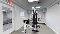 This fitness center offers a clean, efficiently designed workout space suited for strength and resistance training. The room features neutral grey walls and a drop ceiling equipped with flush panel lighting that ensures consistent, bright illumination throughout. A multi-function home gym machine serves as the centerpiece, offering a variety of exercise stations including a lat pulldown, leg curl, and press functions, all finished in a sleek black-and-white design. A standalone leg extension/curl bench complements the main unit, expanding the range of available lower-body exercises. The flooring consists of a low-pile grey carpet with a protective rubber mat placed beneath the equipment to minimize wear and provide added stability during workouts. Two windows along the walls allow for natural light to supplement the overhead fixtures, contributing to an open and welcoming atmosphere despite the room's compact footprint. A wall-mounted vent ensures adequate air circulation, and a door provides straightforward access to and from the space. The overall aesthetic is tidy and functional, with a minimalist approach that keeps the focus on the available equipment.